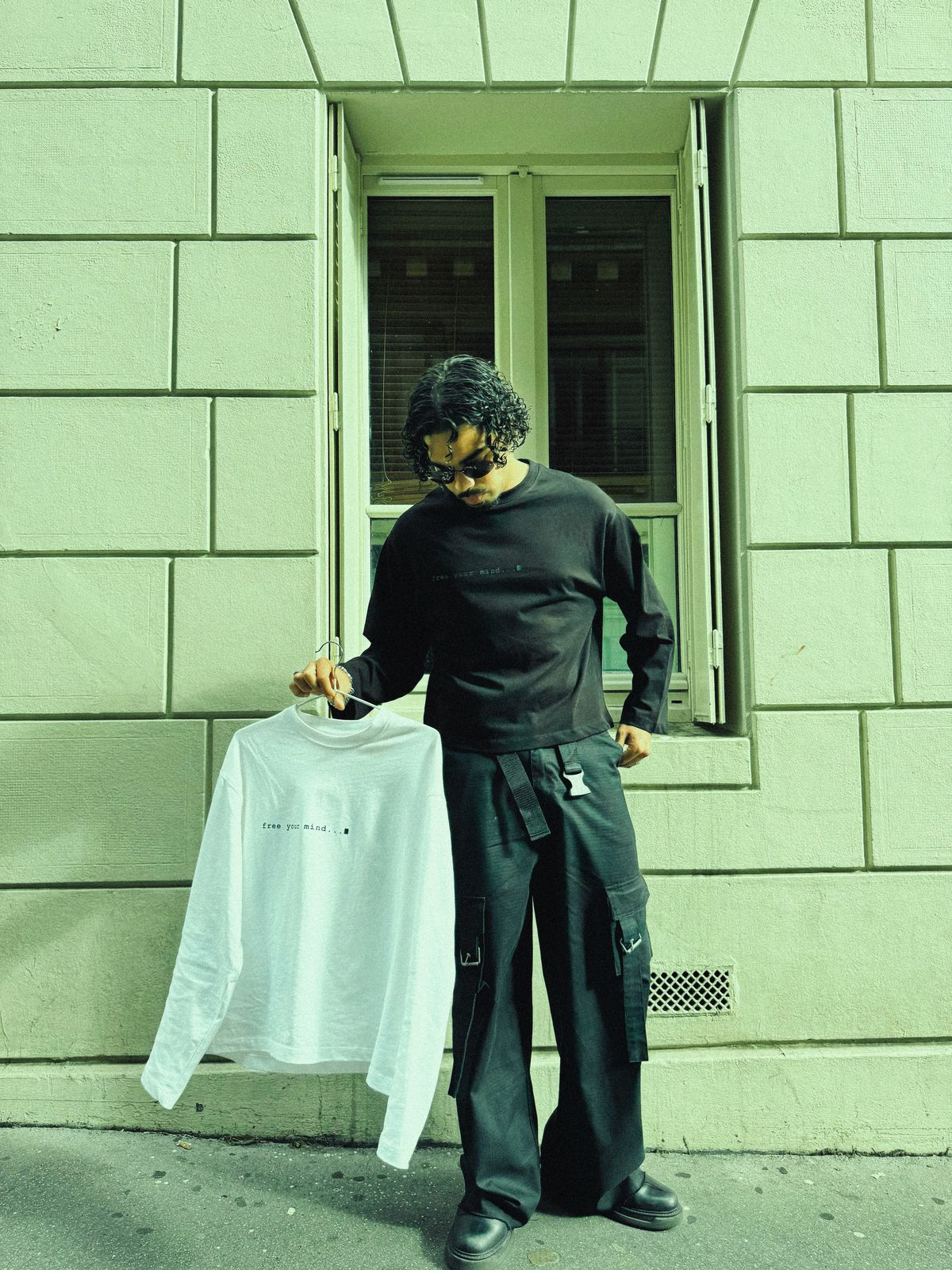Donovan en t-shirt manches longues noir, tenant le modèle blanc sur un cintre devant une façade parisienne.