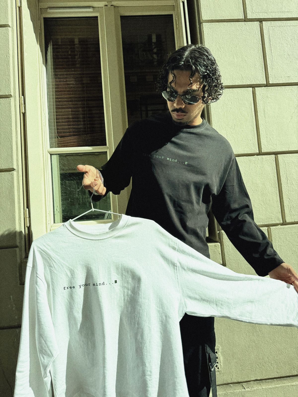 Donovan en t-shirt manches longues noir, présentant le modèle blanc bras tendu devant une fenêtre.