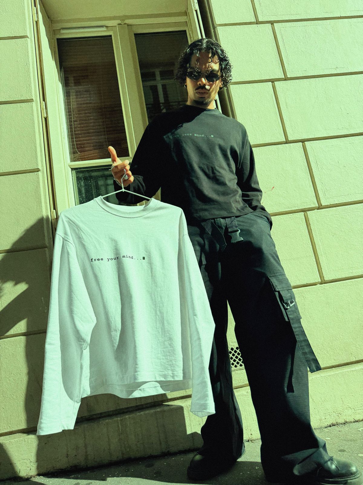 Donovan en t-shirt manches longues noir, le modèle blanc sur un cintre levé devant un mur en pierre.