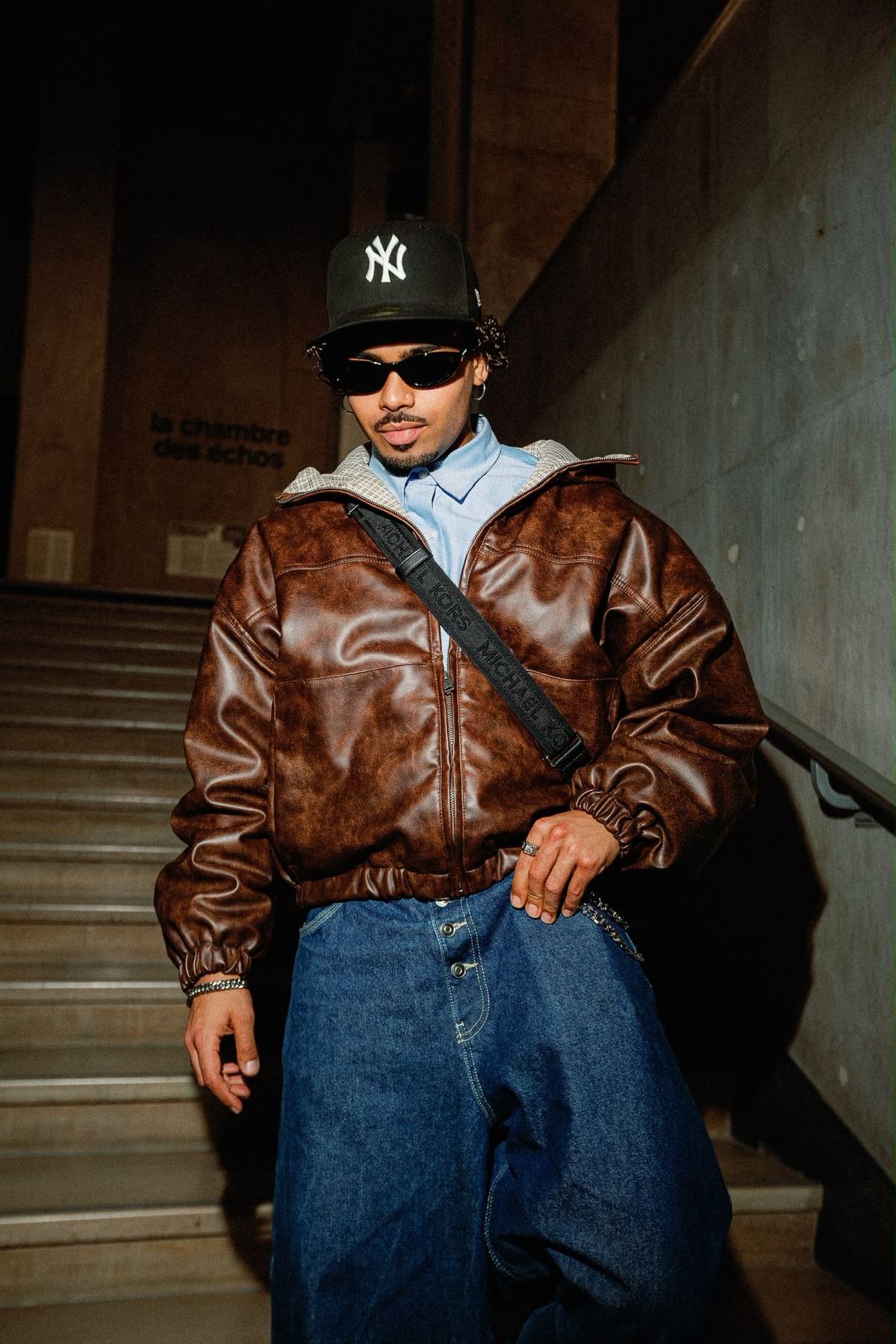 Donovan en blouson de cuir marron et casquette, dans un escalier.