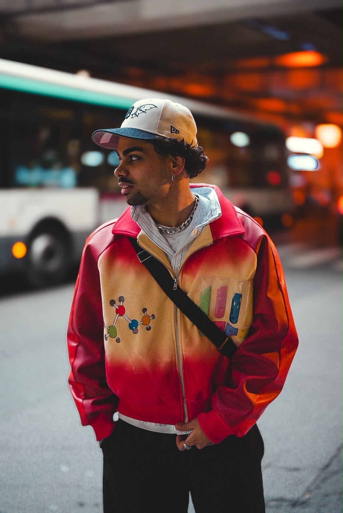 Donovan en blouson dégradé rouge et orange, casquette, devant un bus en ville.