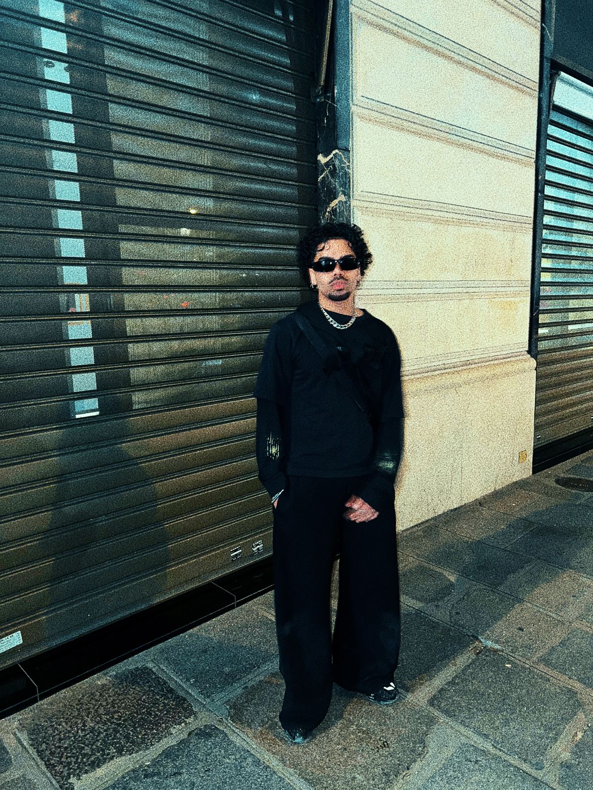 Donovan en lunettes noires, adossé à un rideau métallique dans une rue parisienne.